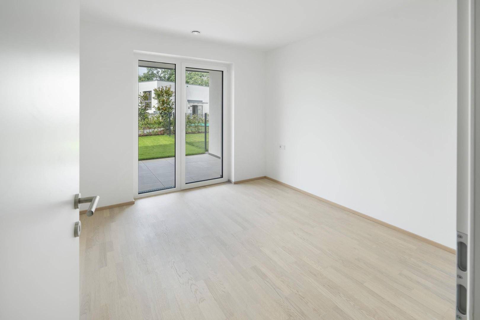 Geräumiges Zimmer mit hellem Holzboden und großem Fenster zum privaten Gartenbereich.