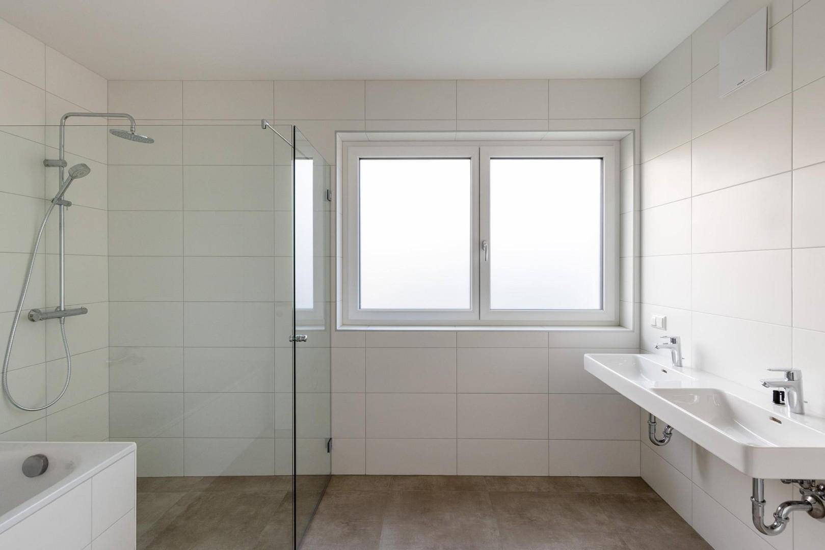 Modernes Badezimmer mit weißen Fliesen, begehbarer Dusche, Badewanne, Doppelwaschbecken und Fenster.