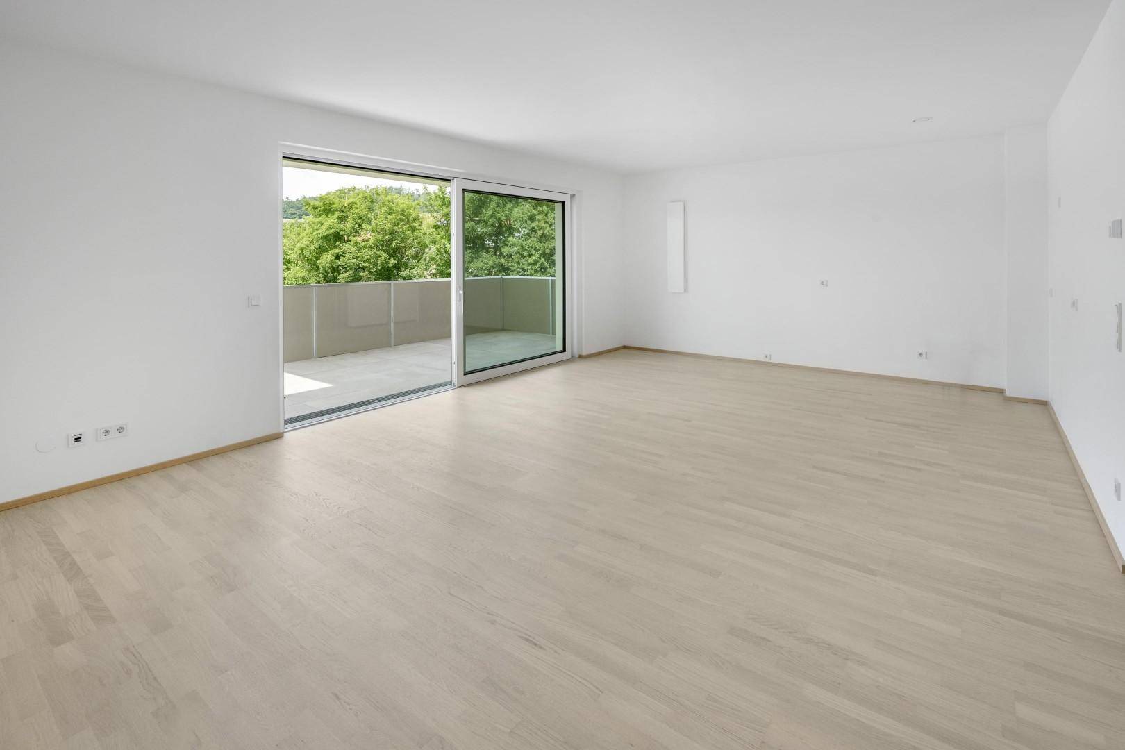 Großzügiges, leeres Wohnzimmer mit hellem Parkettboden und Zugang zu einem großen Balkon.