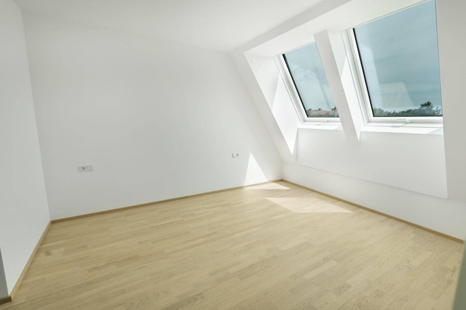 Helles Zimmer im Dachgeschoss mit schrägen Wänden, Holzparkett und zwei Dachfenstern.