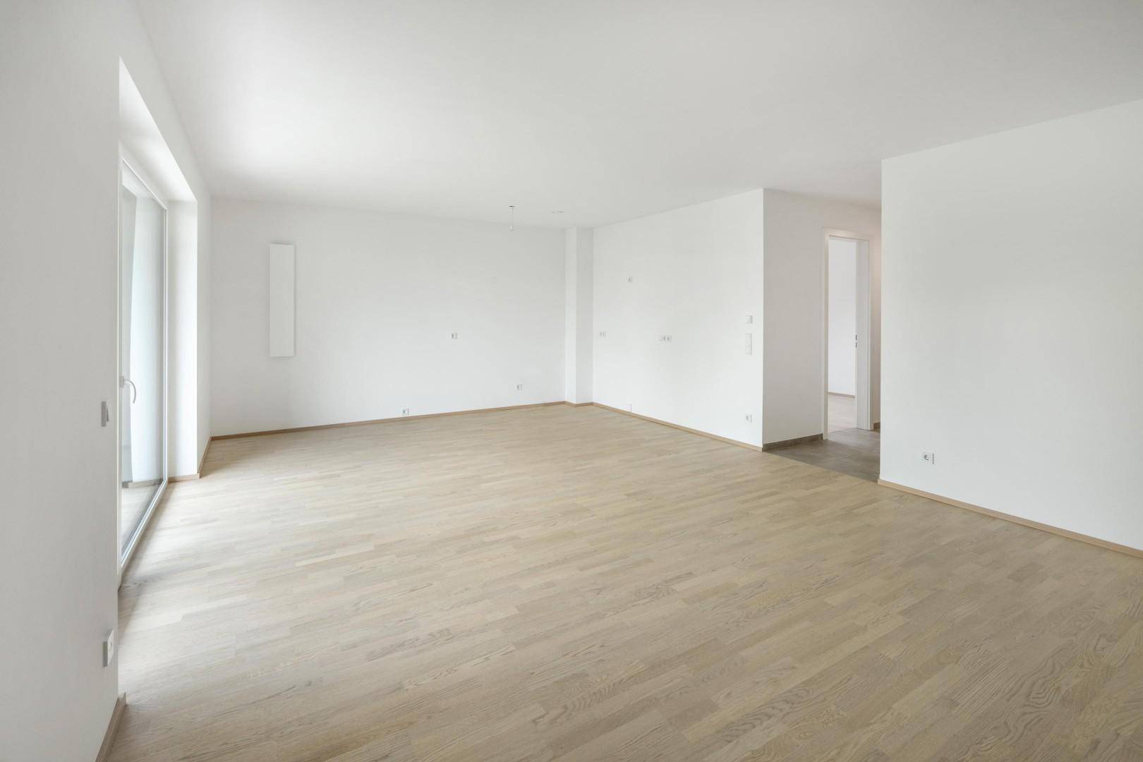 Helles, leeres Wohnzimmer mit großen Fenstern und hellem Holzparkettboden.