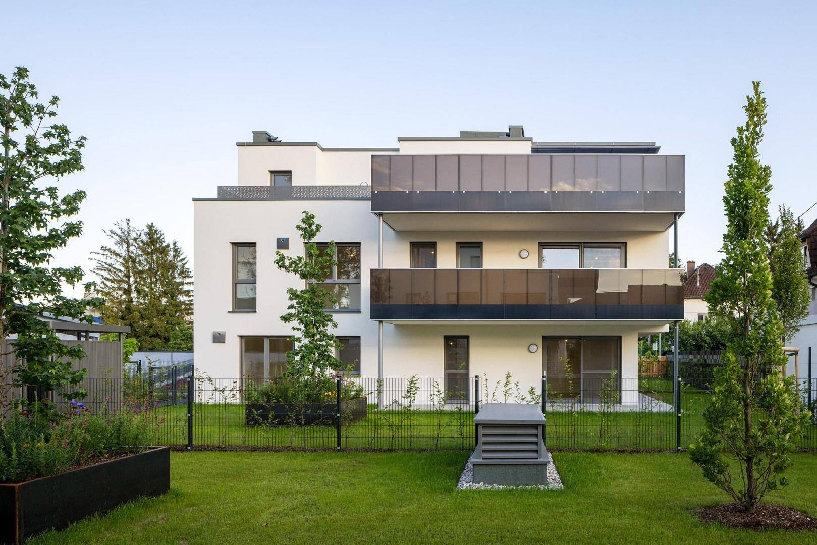 Vorderansicht des modernen Wohnhauses mit Balkonen, Glasgeländern und einem gepflegten Vorgarten.