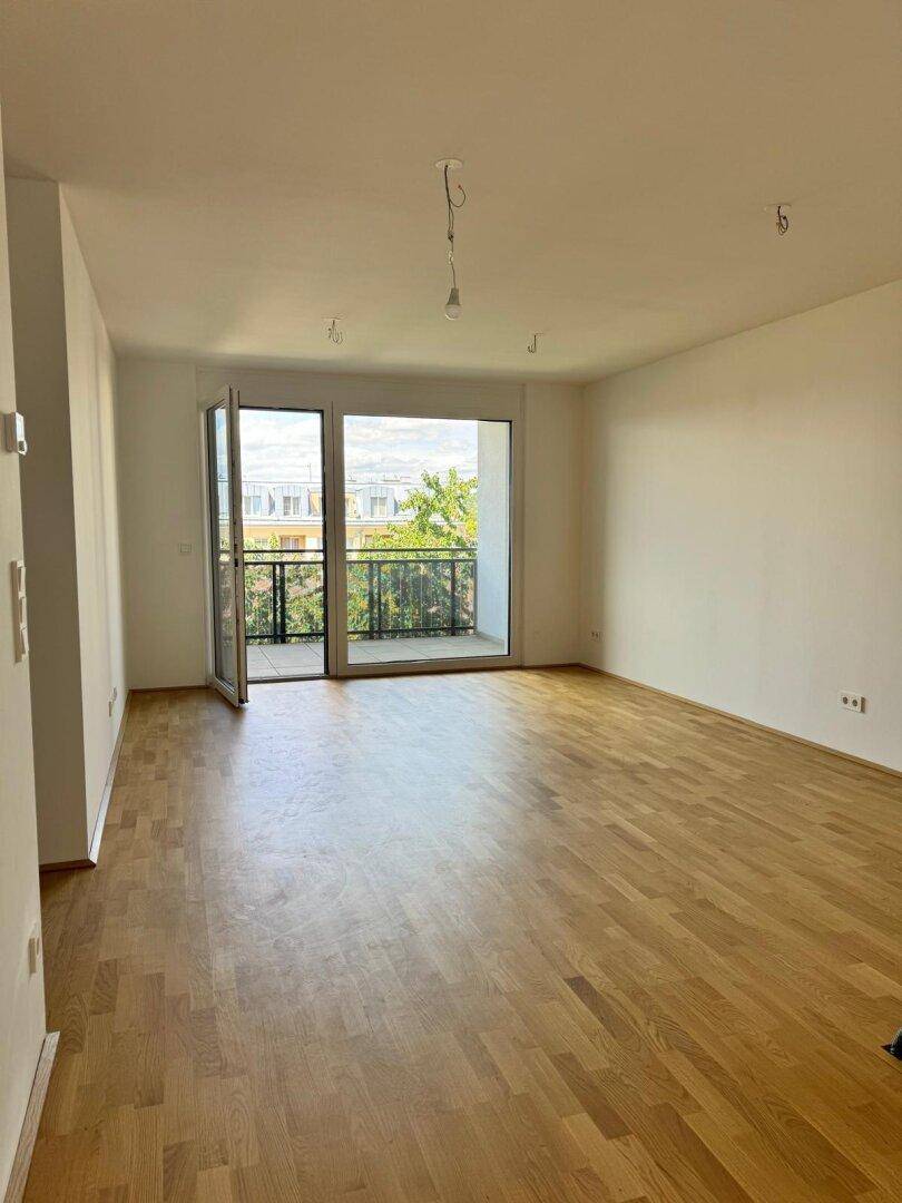 Geräumiges Wohnzimmer mit hellem Parkettboden und offener Balkontür, die viel Tageslicht hereinlässt.