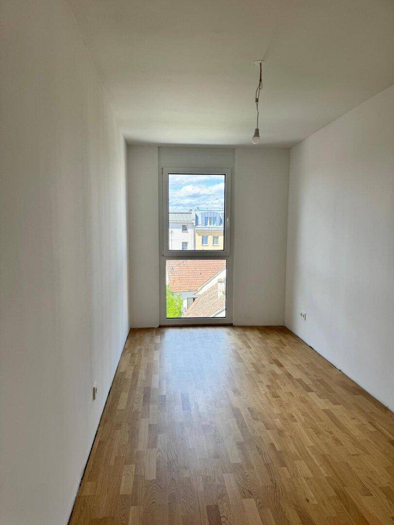 Leeres Schlafzimmer mit Holzparkettboden und einem hohen Fenster, das einen Blick auf die Stadt bietet.