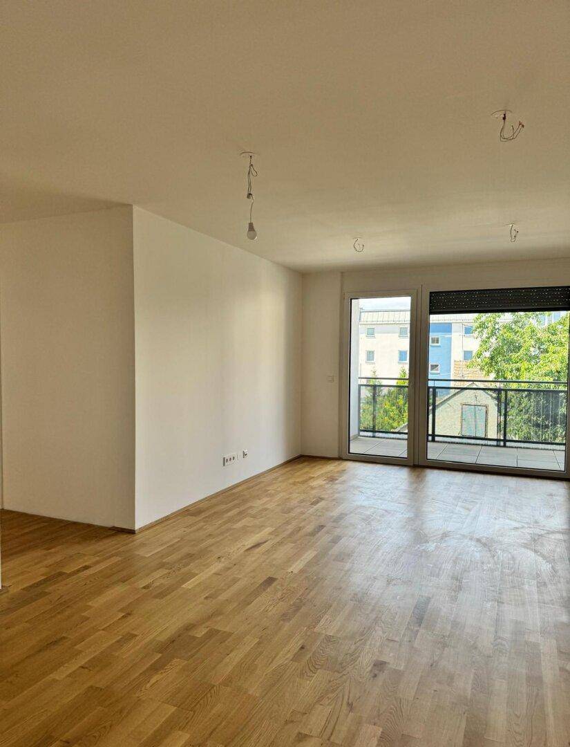 Helles, leeres Wohnzimmer mit Parkettboden und großen Fenstern, die Zugang zu einem Balkon bieten.
