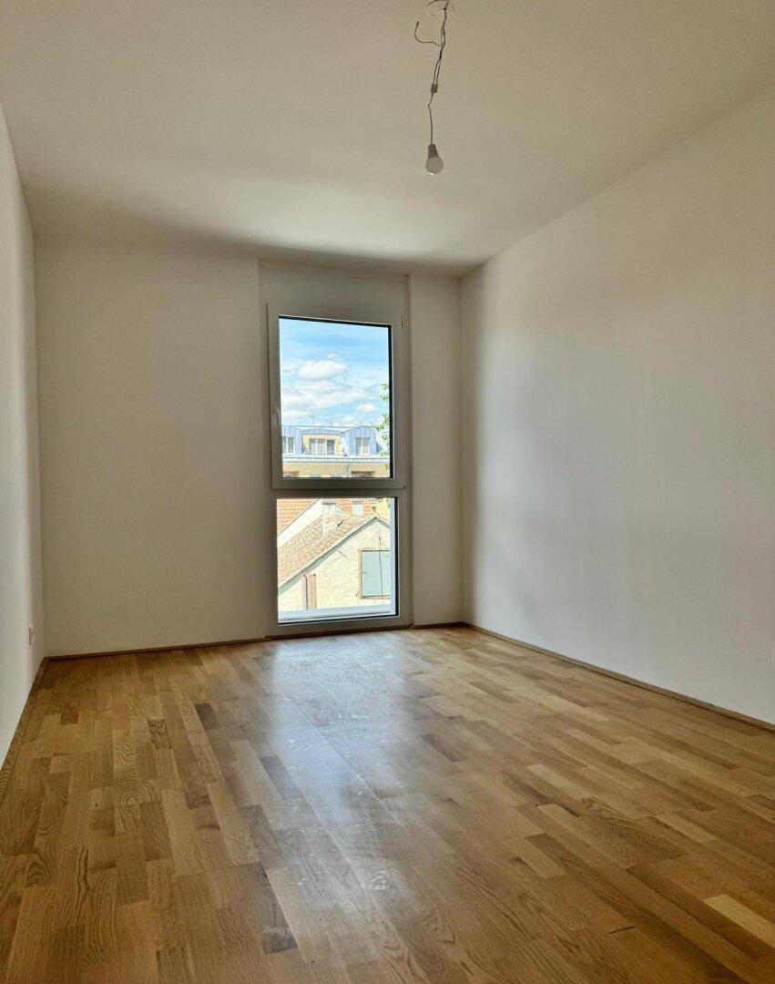Leeres Schlafzimmer mit Holzparkettboden und einem hohen Fenster, das einen Blick auf die Stadt bietet.
