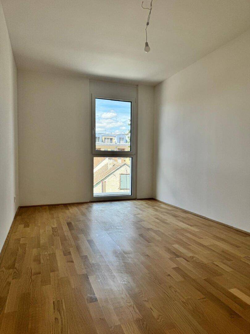 Leeres Schlafzimmer mit hellem Parkettboden und einem großen Fenster, das viel Tageslicht hereinlässt.