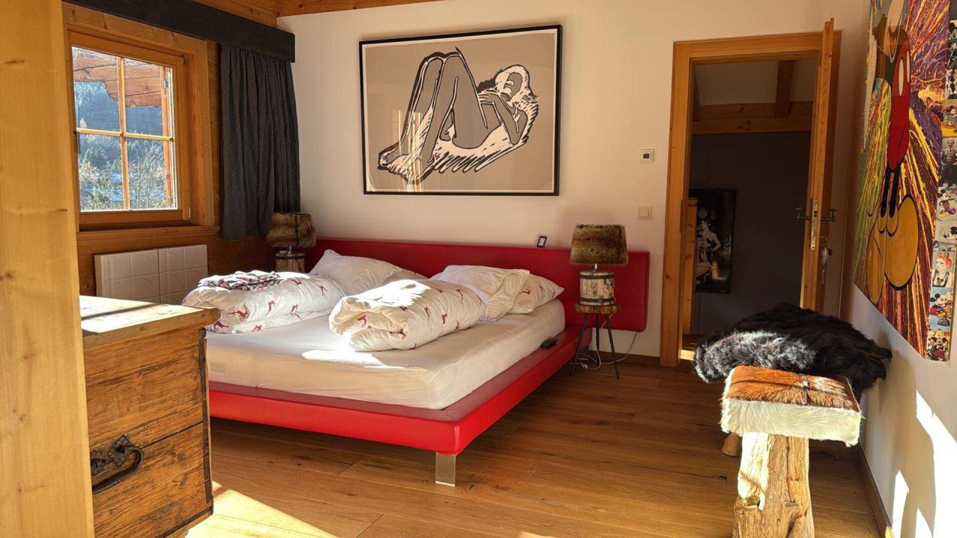 Modernes Schlafzimmer mit rotem Bett, Holzboden und viel natürlichem Licht.