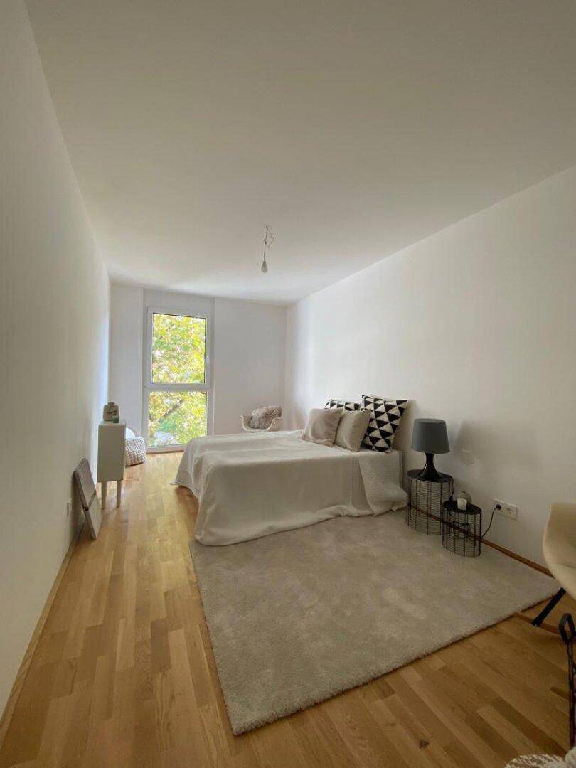 Helles Schlafzimmer mit Holzboden, weiß gestrichenen Wänden und großem Fenster für viel Tageslicht.