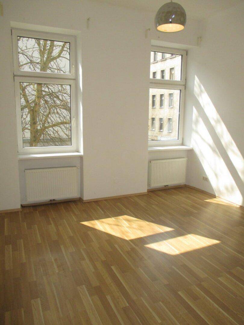 Lichtdurchflutetes Zimmer mit Laminatboden, weißen Wänden und Blick auf Bäume und ein Gebäude.