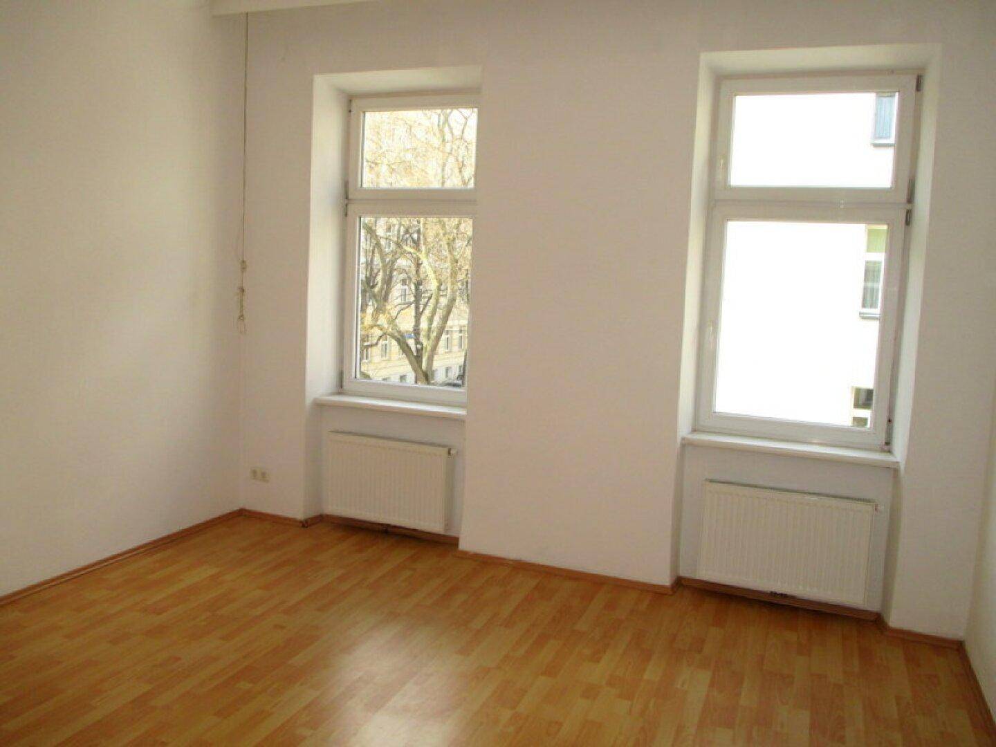 Geräumiges Zimmer mit hellem Laminatboden, weißen Wänden und zwei Fenstern mit Blick ins Grüne.