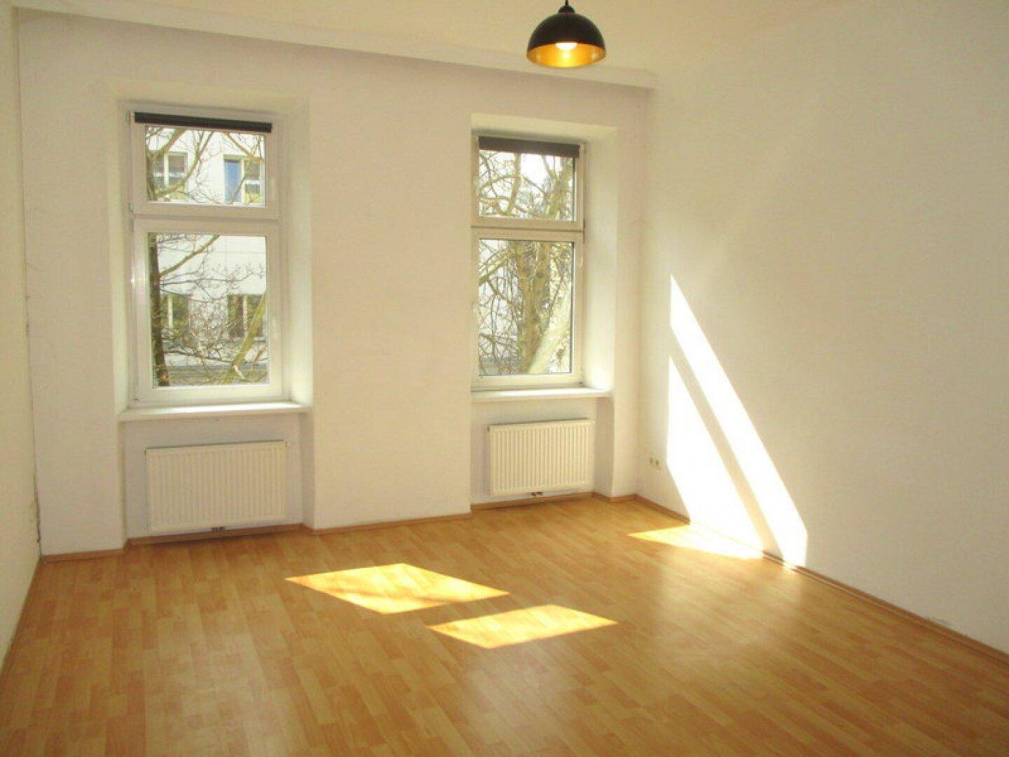 Freundliches Zimmer mit Laminatboden, weißen Wänden und viel natürlichem Licht durch die großen Fenster.