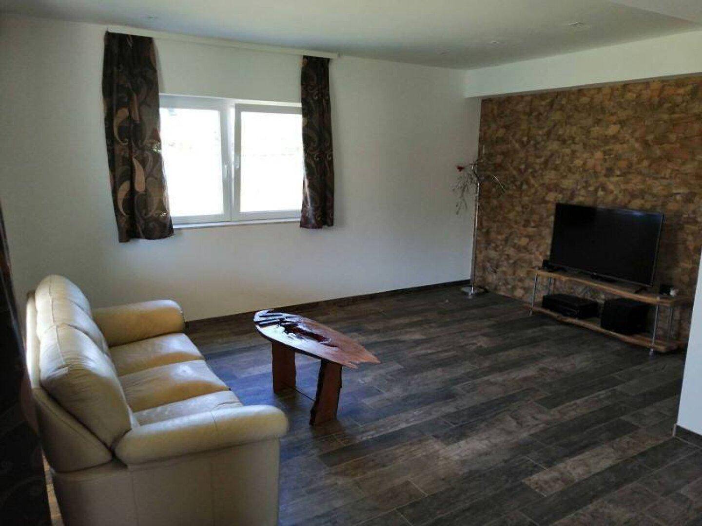 Gemütliches Wohnzimmer mit Holzoptik-Fliesen, beiger Couch und einer Wandverkleidung aus Naturstein.