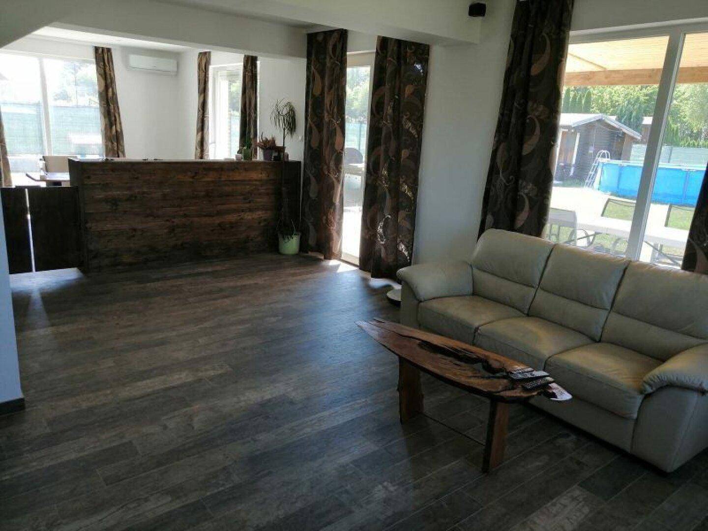 Heller Wohnbereich mit Holzoptik-Fliesen, beiger Couch und Blick auf den Garten und Pool.