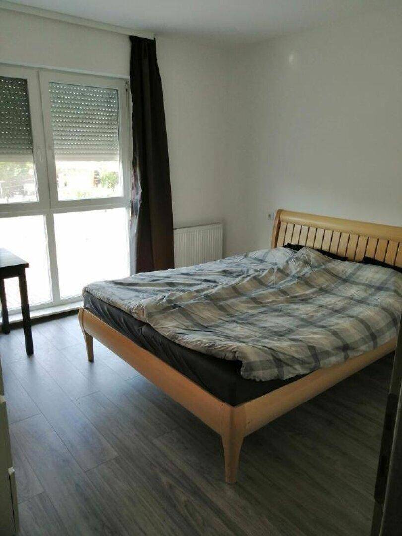 Helles Schlafzimmer mit Holzbett, Holzoptik-Fliesen und großen Fenstern mit Rollläden.