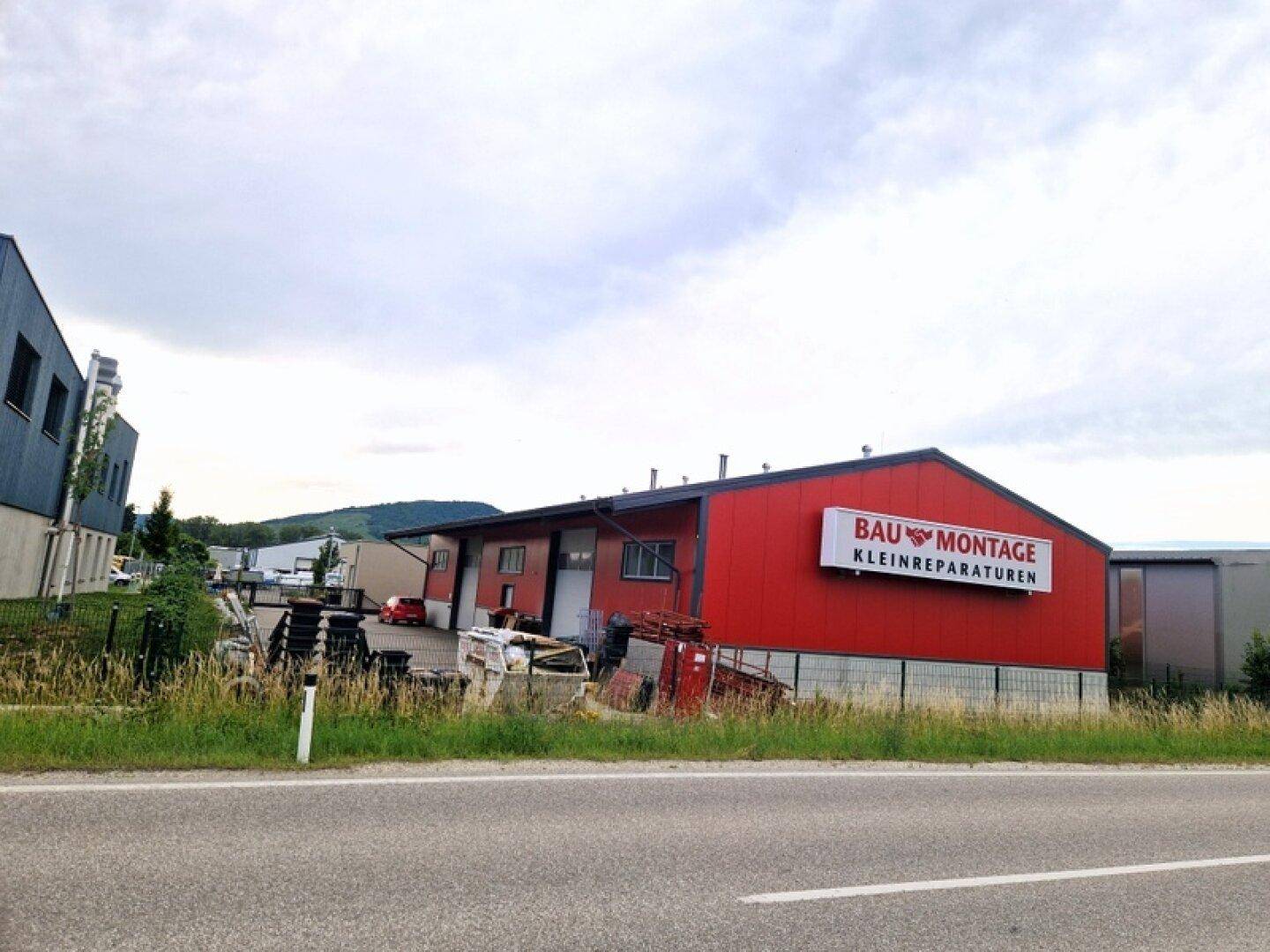 Rotes Industriegebäude mit Beschriftung und umzäuntem Gelände, von der Straße aus gesehen.