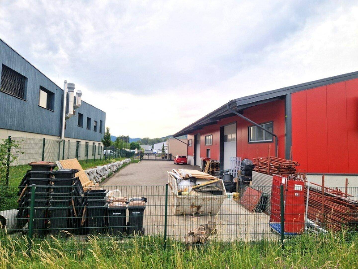 Blick auf zwei Industriegebäude mit einem asphaltierten Zufahrtsweg und gelagerten Materialien.