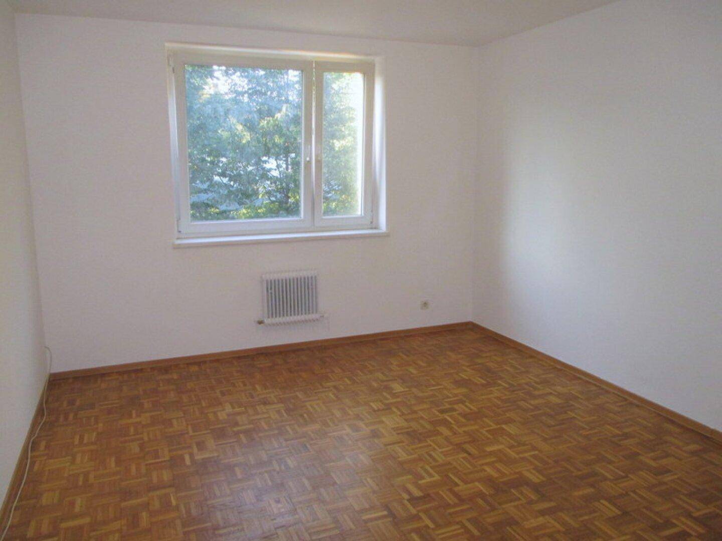 Geräumiges Zimmer mit Parkettboden und großem Fenster, das viel Tageslicht hereinlässt.