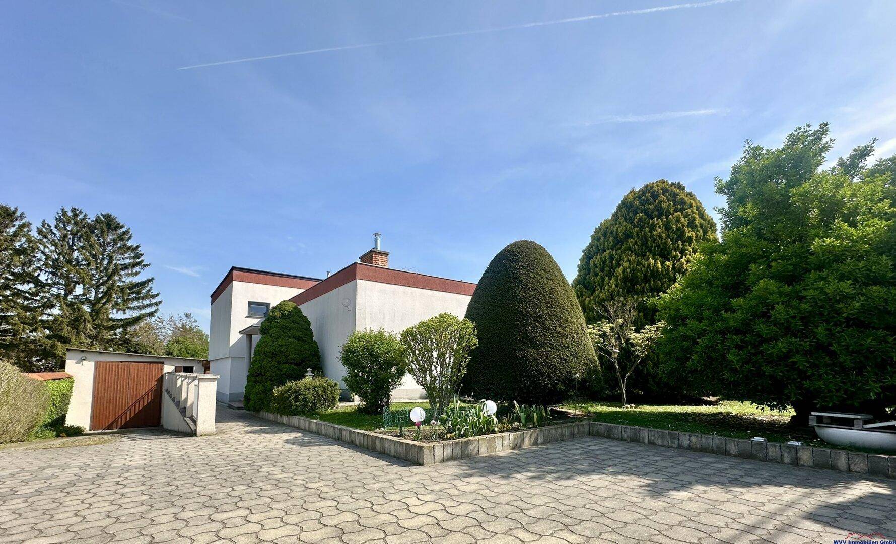 Ansicht des Hauses mit gepflasterter Einfahrt und gepflegten Sträuchern unter blauem Himmel.