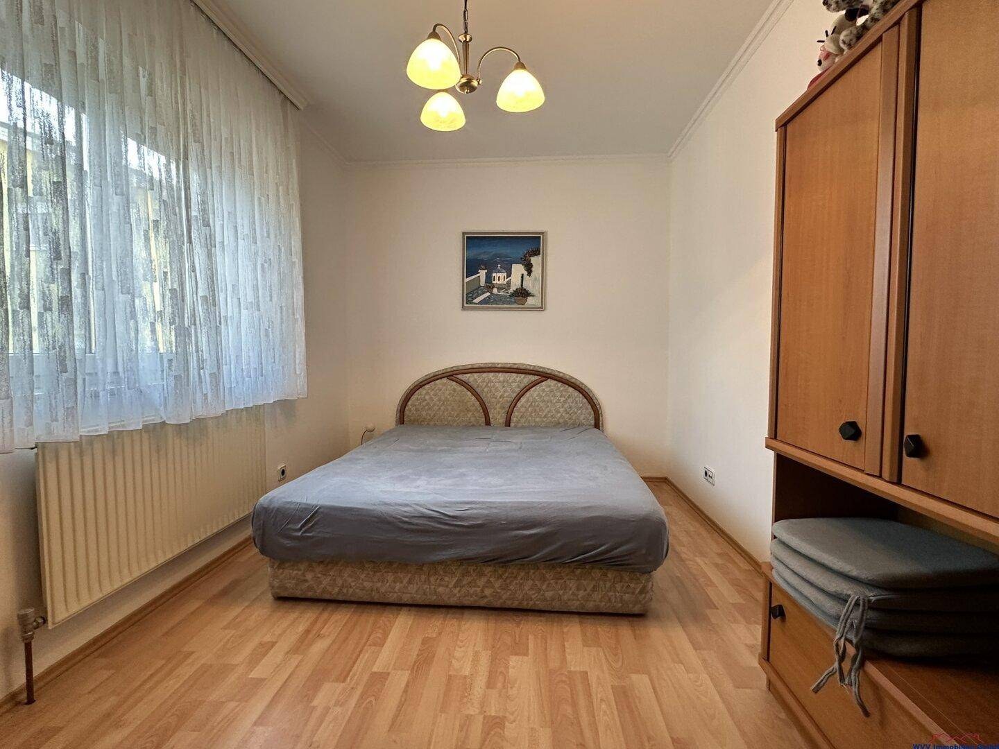 Helles Schlafzimmer mit Laminatboden, großem Fenster und schlichtem Bett.