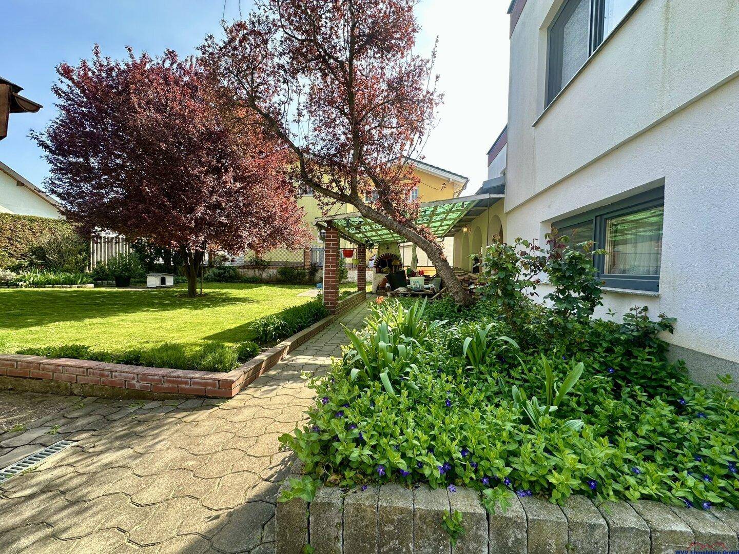 Seitlicher Blick auf den Garten mit blühenden Pflanzen, gepflastertem Weg und überdachtem Bereich.