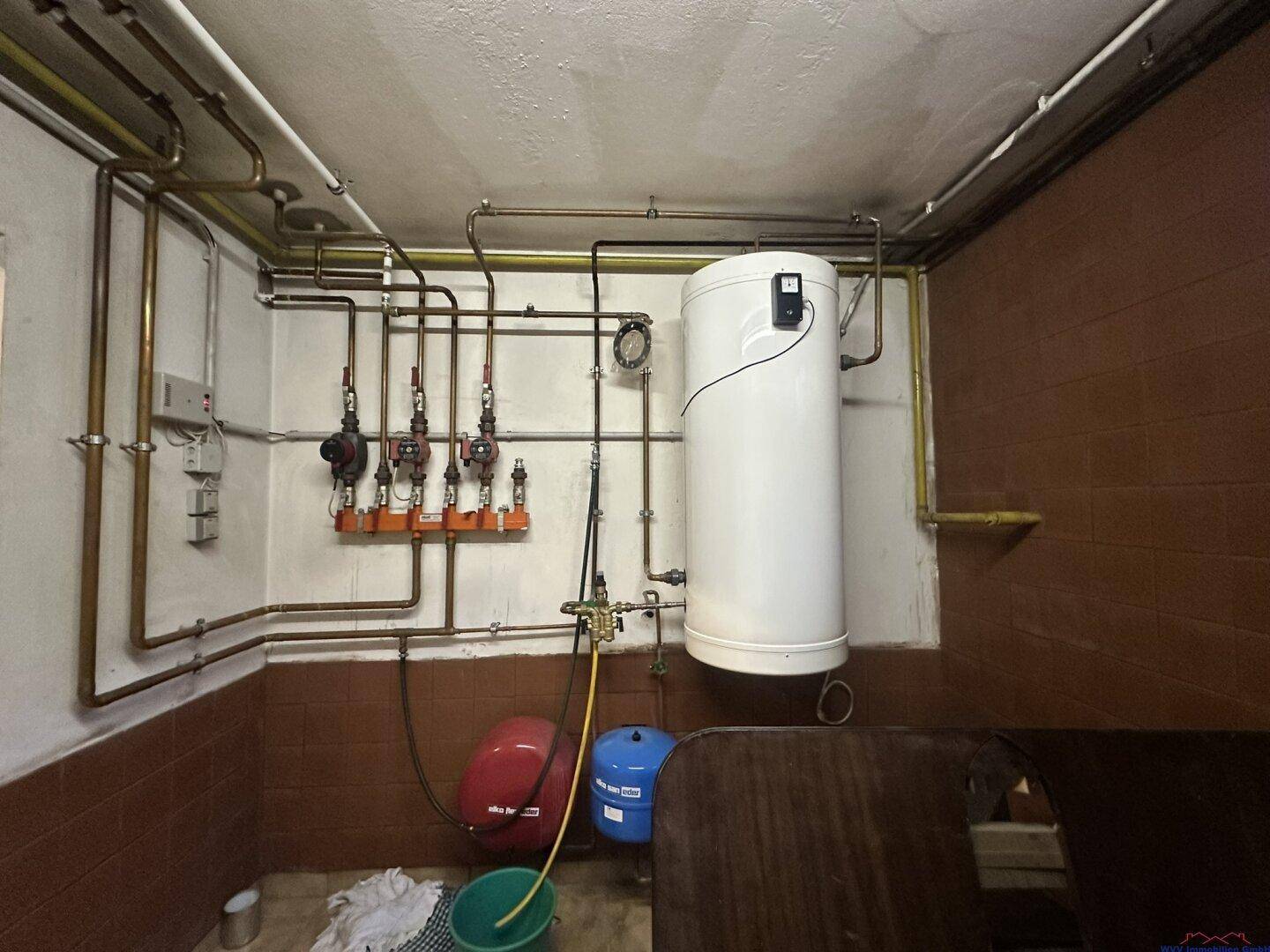 Heizungsraum im Keller mit Warmwasserspeicher und sichtbaren Rohrleitungen.