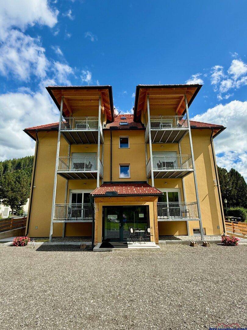 Frontansicht eines modernen Apartmenthauses mit gelber Fassade, großen Fenstern und überdachten Balkonen.