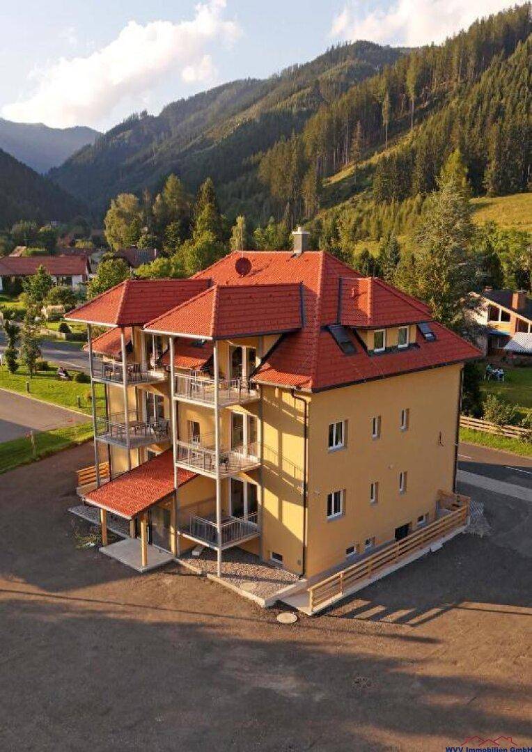 Modernes Apartmenthaus mit mehreren Balkonen und rotem Ziegeldach in alpiner Berglandschaft.