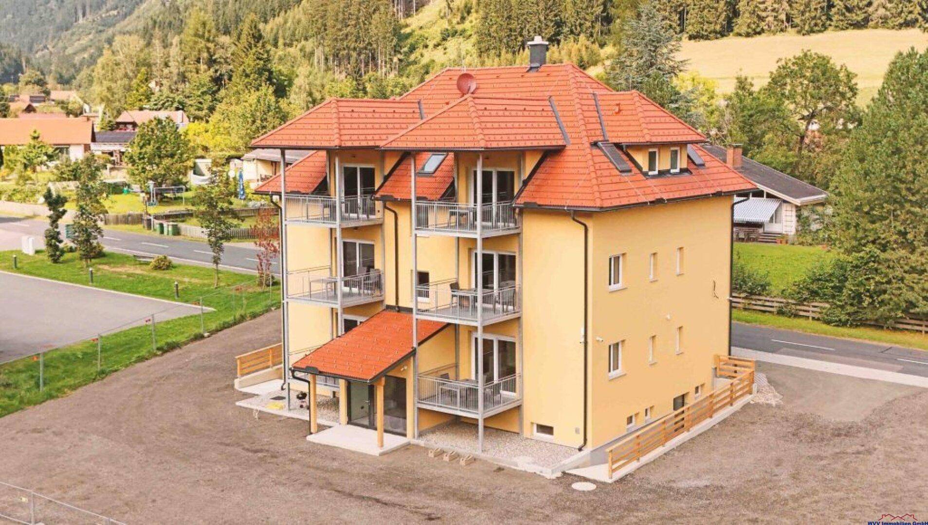 Neubau mit gelber Fassade, roten Dächern und mehreren Balkonen, umgeben von Natur.