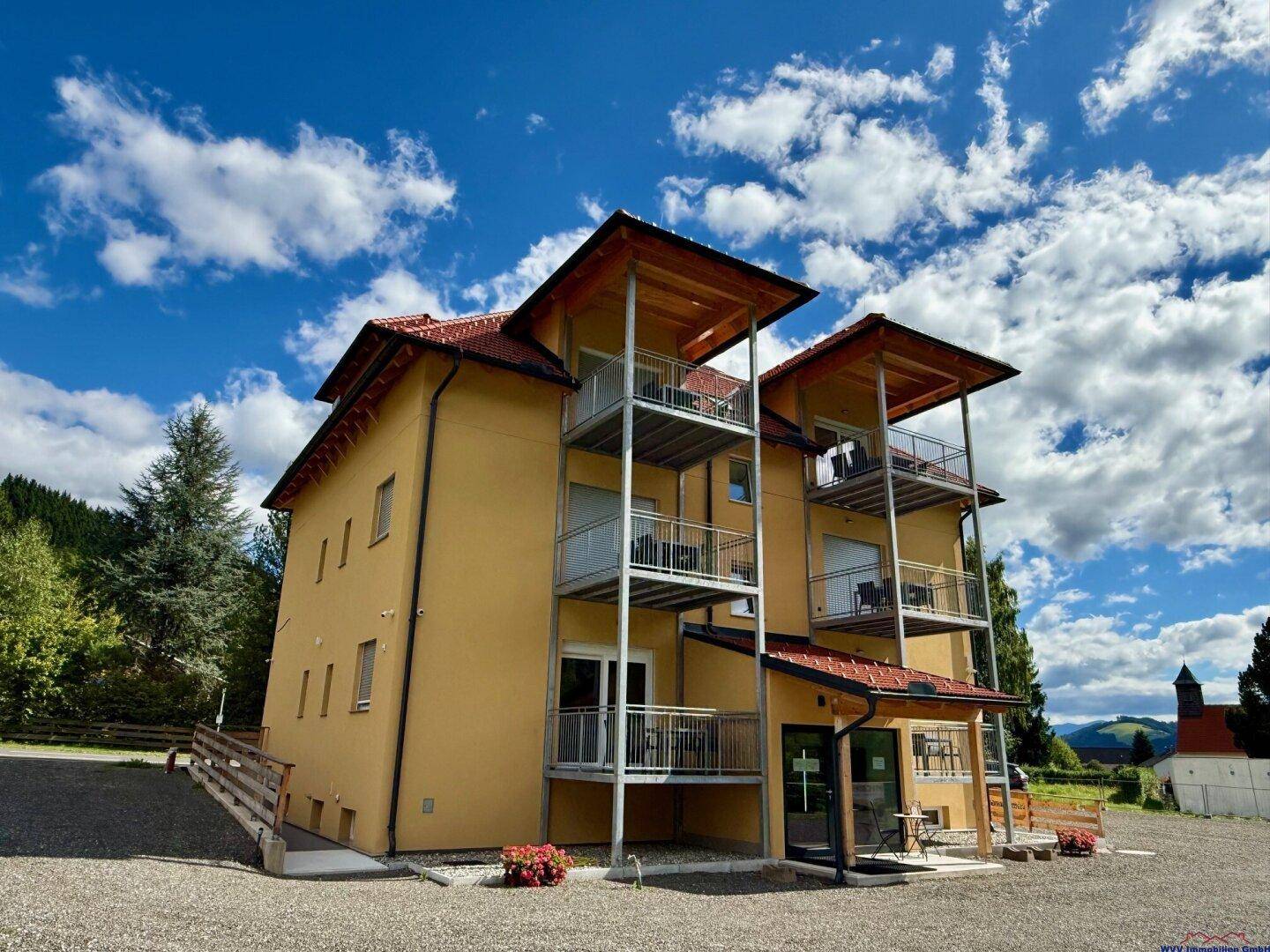 Seitenansicht des modernen Apartmenthauses mit gelber Fassade, Holzbalkonen und Blick auf die Berge.