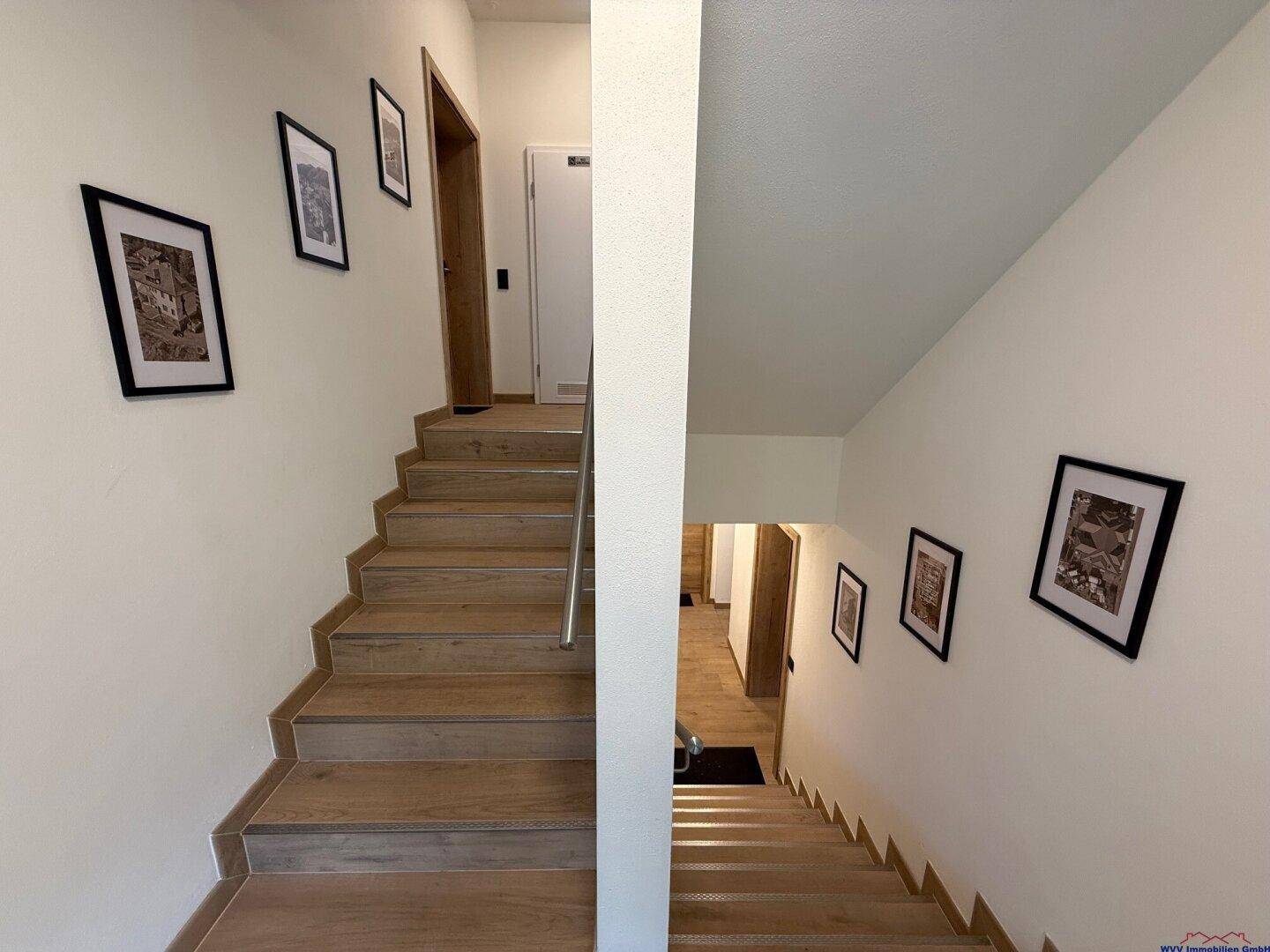 Heller Treppenaufgang mit Holzstufen und weißen Wänden, geschmückt mit gerahmten Bildern.