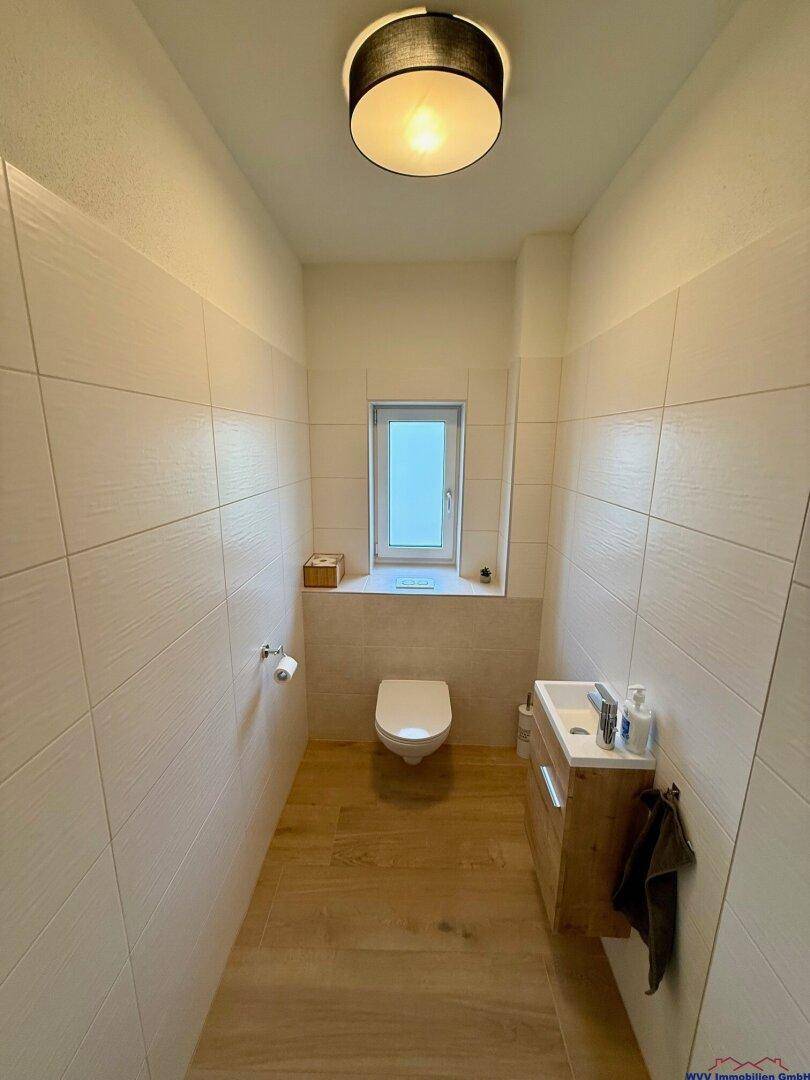 Separates WC mit wandhängender Toilette, kleinem Waschbecken und Fenster.