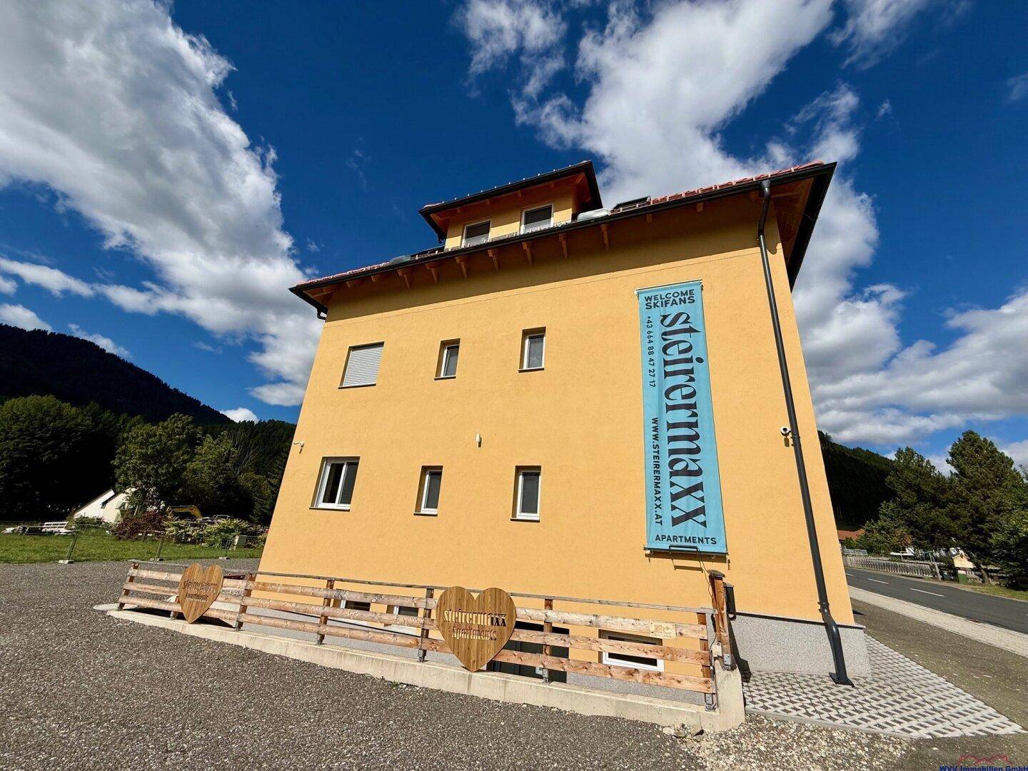 Seitenansicht des Apartmenthauses mit gelber Fassade und Werbebanner, umgeben von ländlicher Natur.