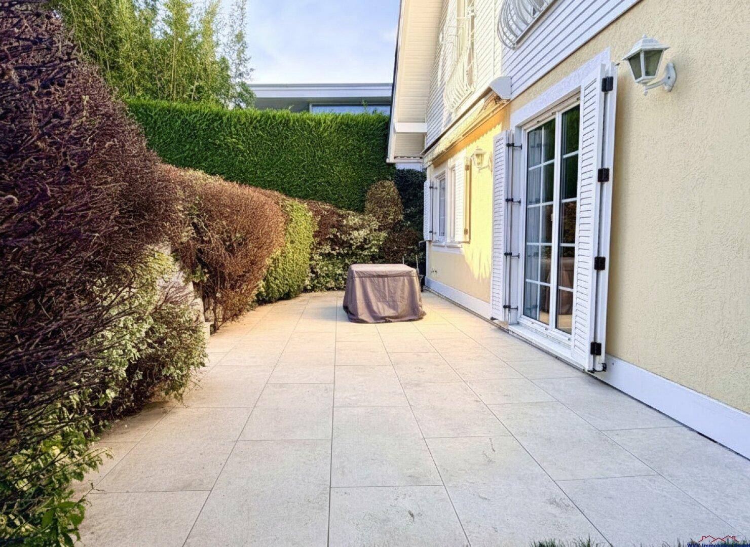 Sonnige Terrasse mit Fliesenboden und Zugang zum Garten, umgeben von Hecken.