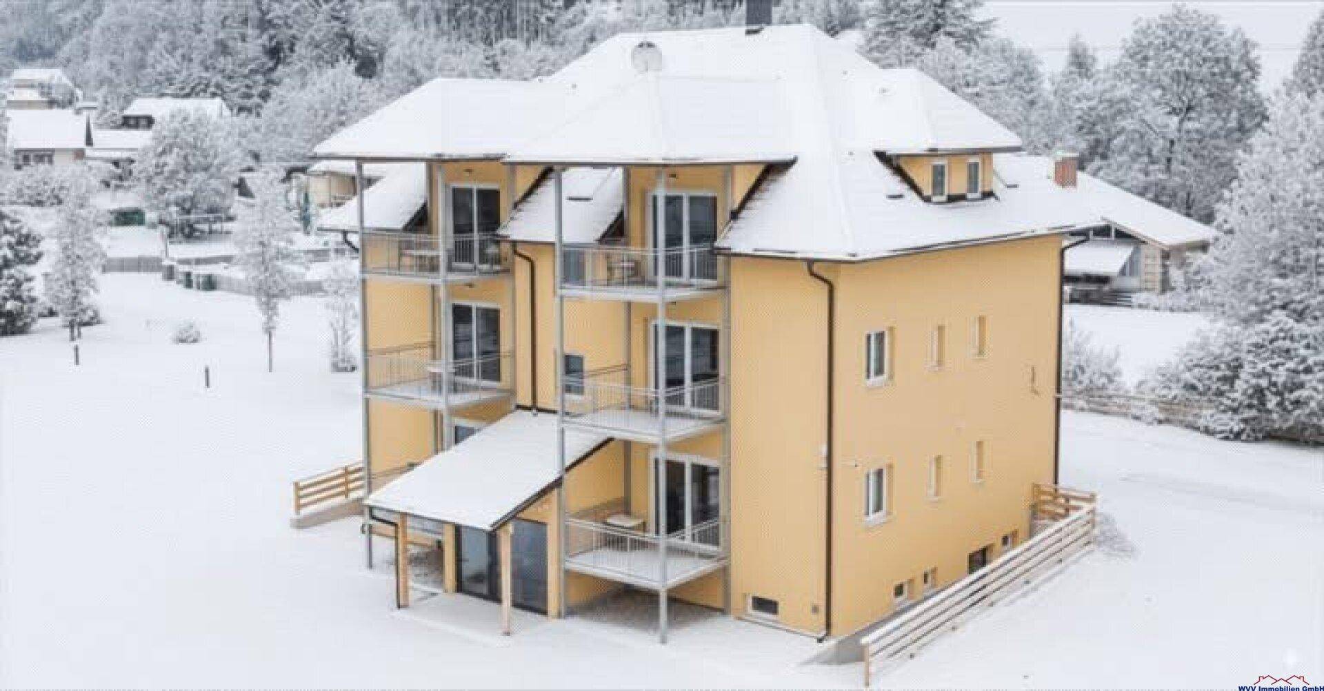 Modernes Mehrfamilienhaus in winterlicher Landschaft mit mehreren Balkonen und großen Fenstern.