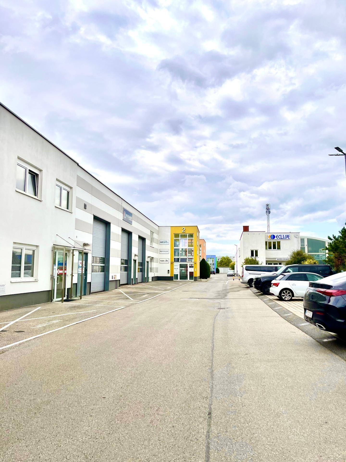 Blick auf eine Gewerbestraße mit mehreren Gebäuden, Parkplätzen und einem bewölkten Himmel.