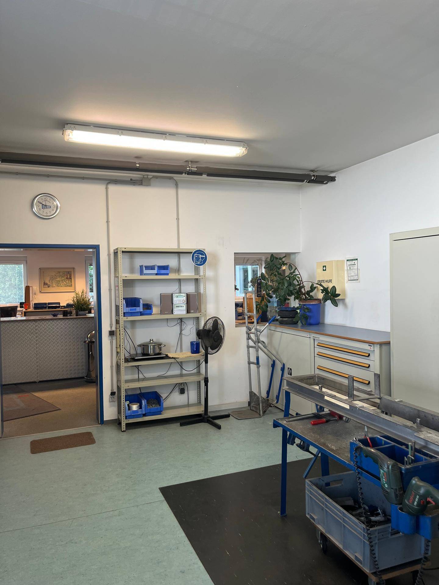 Werkstatt mit Arbeitsbereich, Regalen und einem offenen Durchgang zu einem Empfangsbereich.