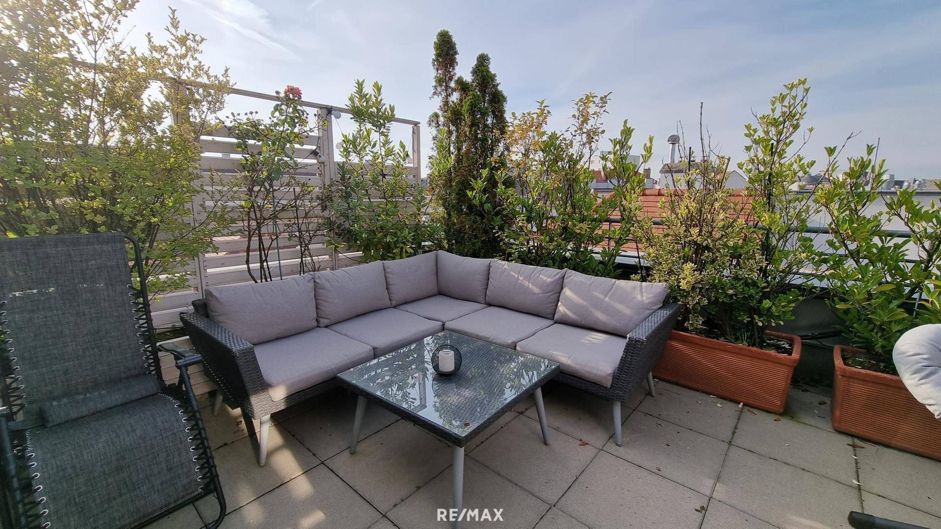 Großzügige Dachterrasse mit bequemer Lounge-Ecke und grüner Bepflanzung unter blauem Himmel.