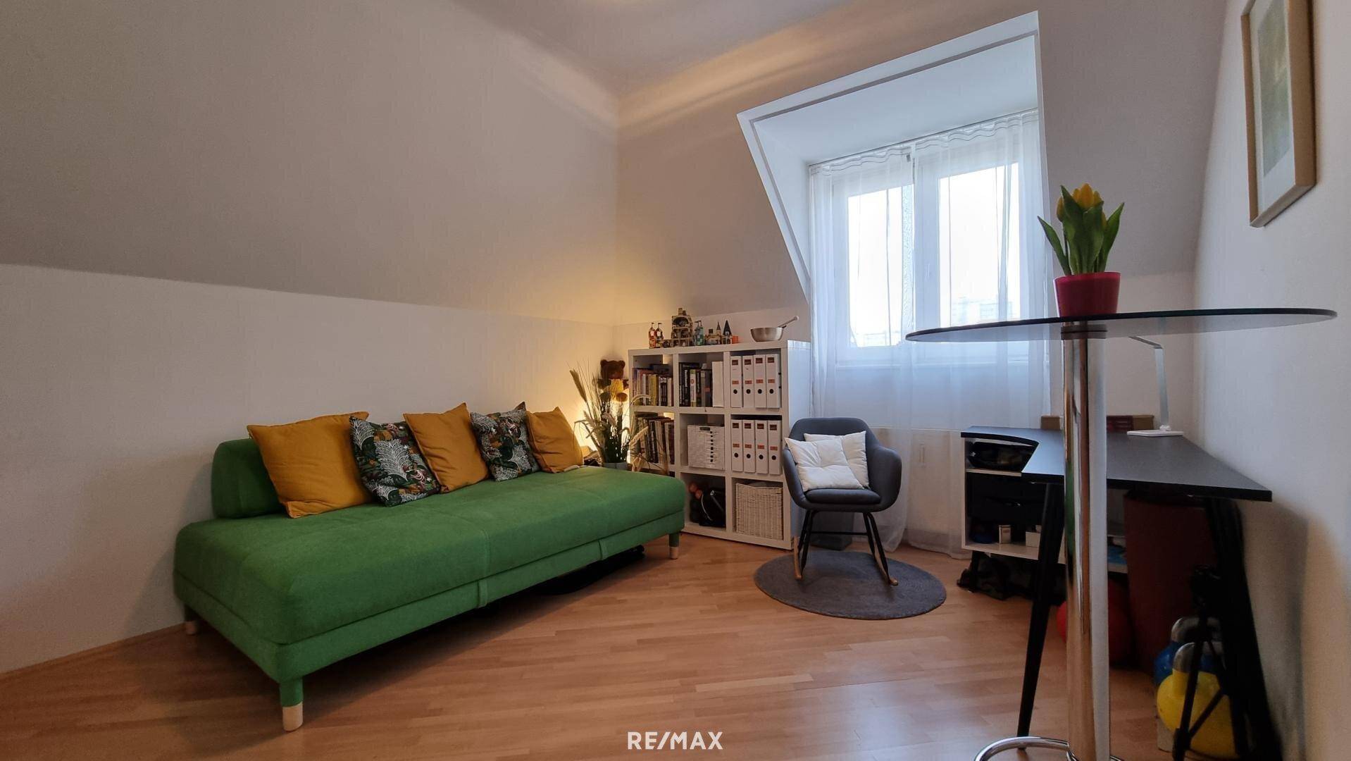 Helles Zimmer mit grünem Sofa, Bücherregal und Schreibtischbereich unter der Dachschräge.