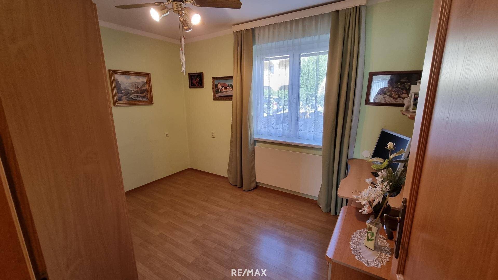 Helles Zimmer mit Laminatboden, Fenster und einfacher Einrichtung.