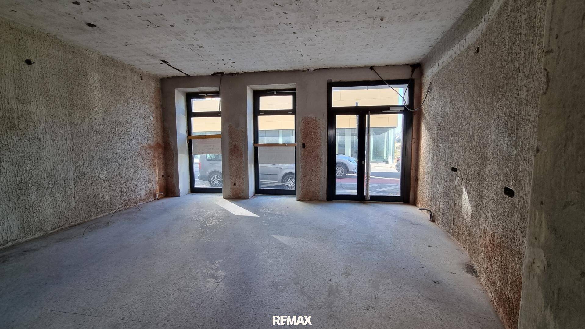 Geräumiger Geschäftsraum im Rohbau mit Blick auf die Straße durch große Fenster.