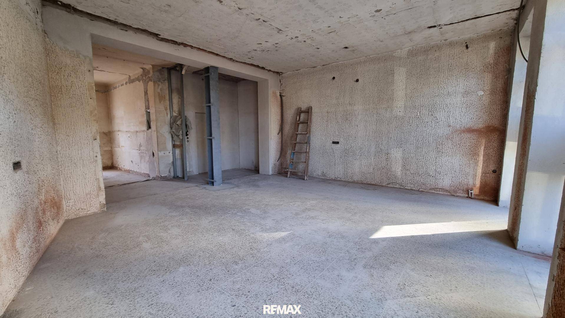 Weiter Geschäftsraum im Rohbau mit unfertigen Wänden und einem Durchgang zu weiteren Bereichen.