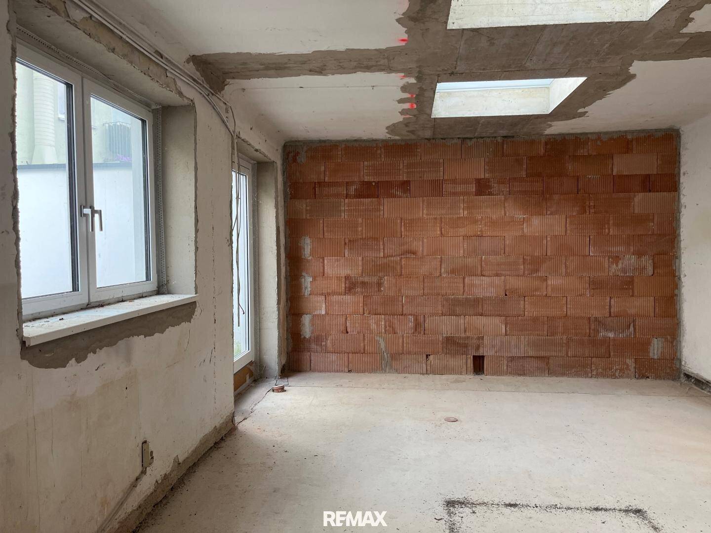 Raum im Rohbau mit unverputzten Ziegelwänden und großen Fenstern, die Tageslicht hereinlassen.