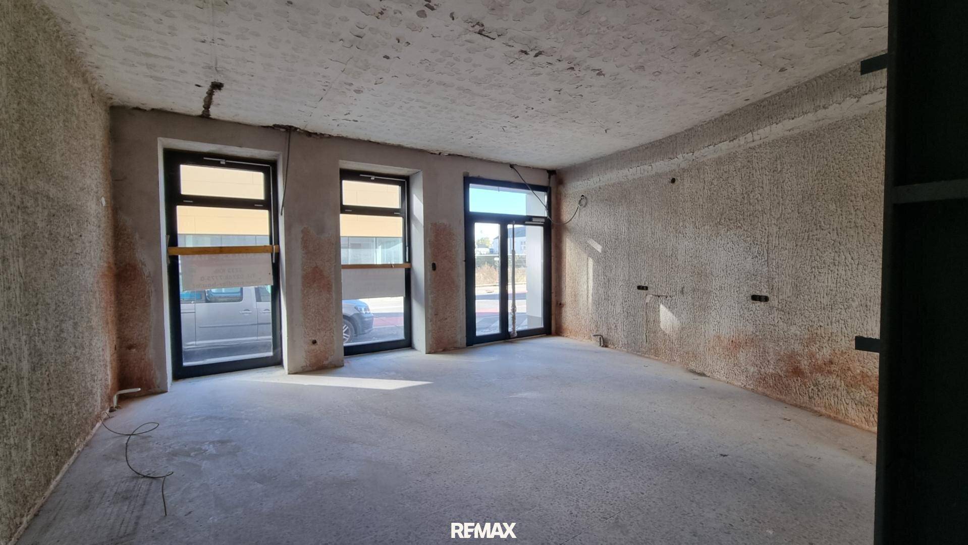 Großer, leerer Geschäftsraum im Rohbau mit mehreren Fenstern zur Straße hin.