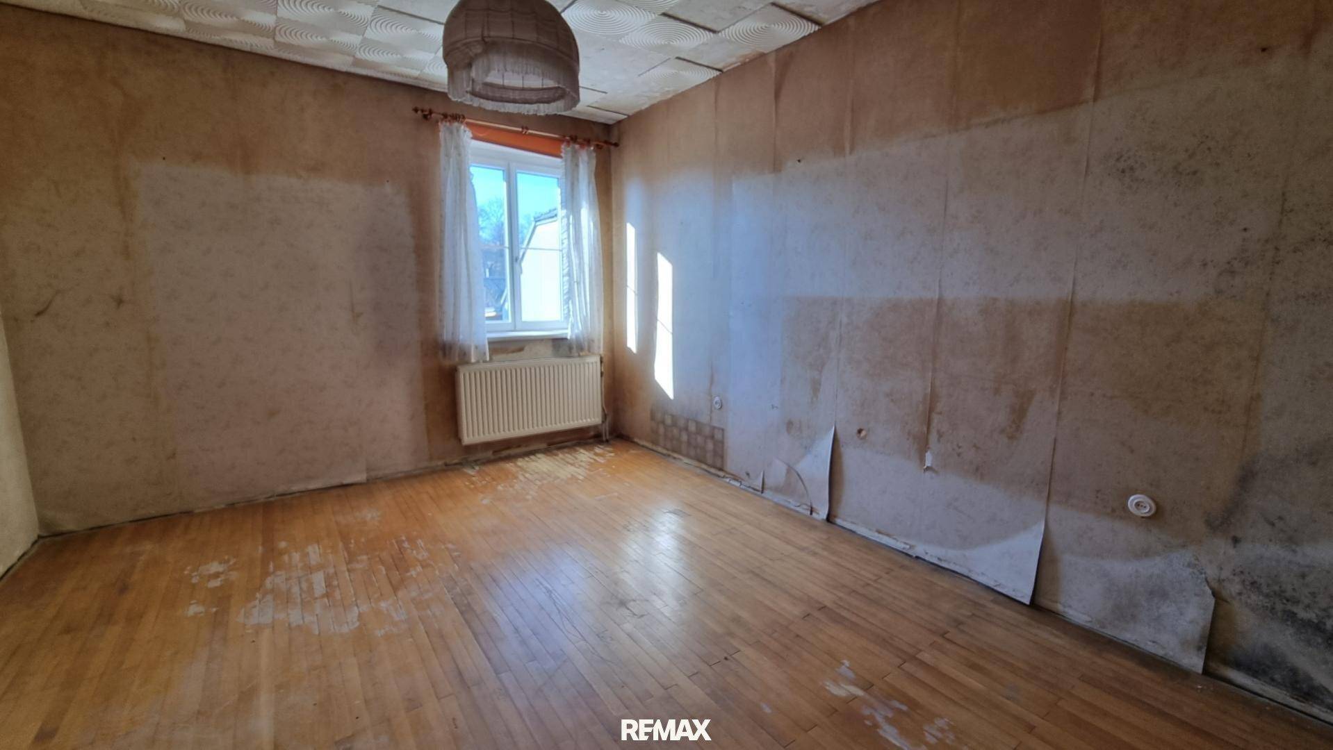 Schlafzimmer mit Holzdielenboden, Tapetenresten und einem Fenster, das viel Sonnenlicht hereinlässt.