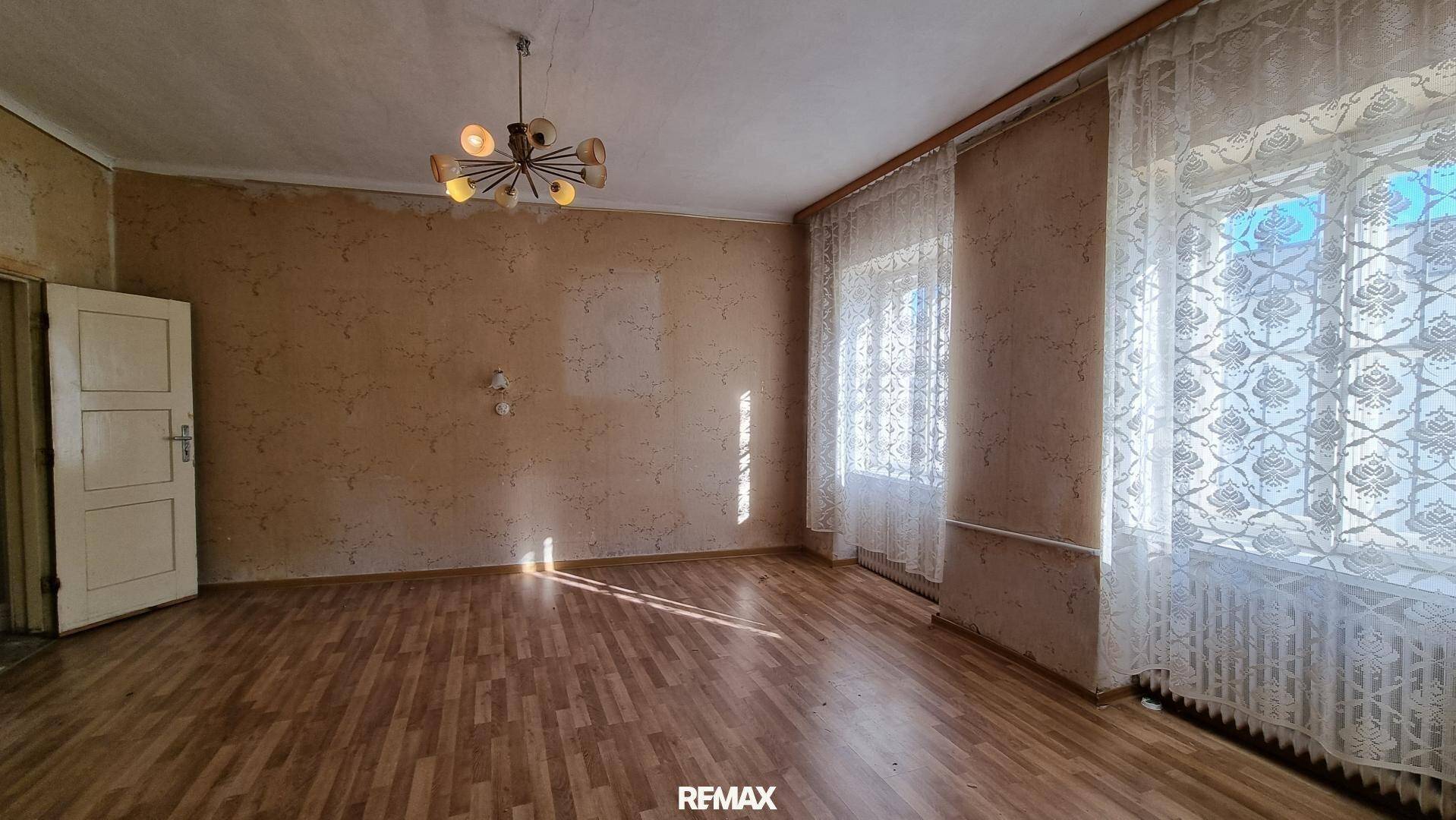 Leeres Wohnzimmer mit Laminatboden, Tapetenresten und zwei großen Fenstern.