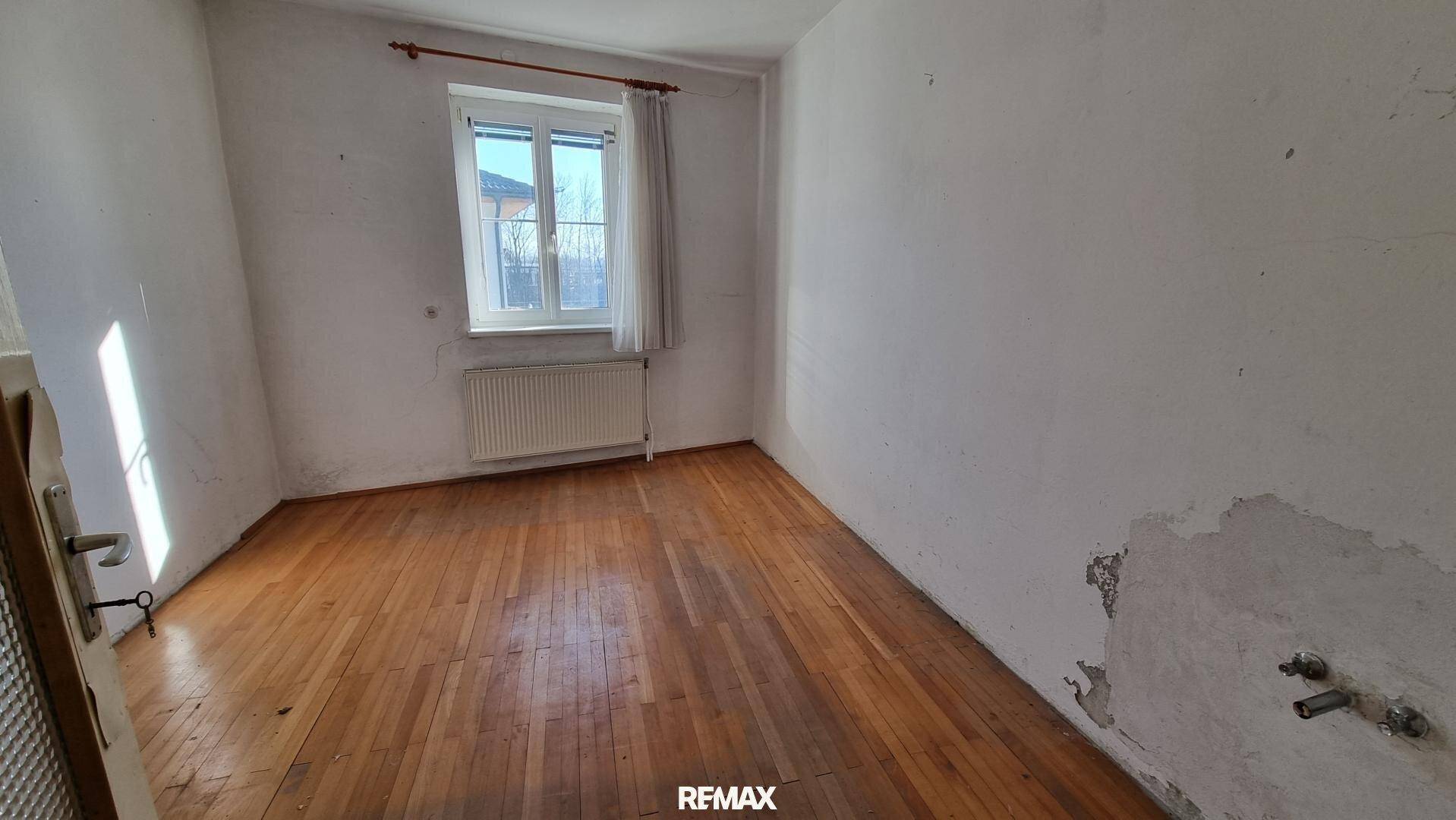 Leeres Schlafzimmer mit Holzdielenboden, Heizkörper und einem Fenster, das Tageslicht spendet.