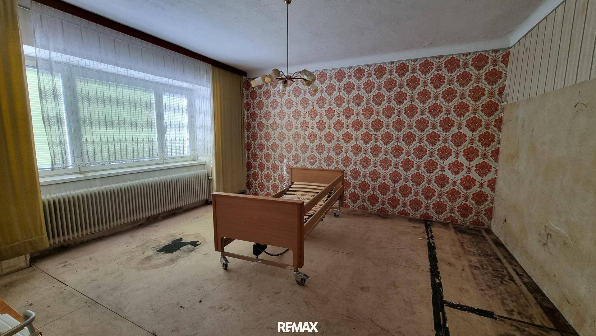 Schlafzimmer mit alter Tapete, einem Einzelbett und einem Heizkörper unter dem Fenster, renovierungsbedürftig.