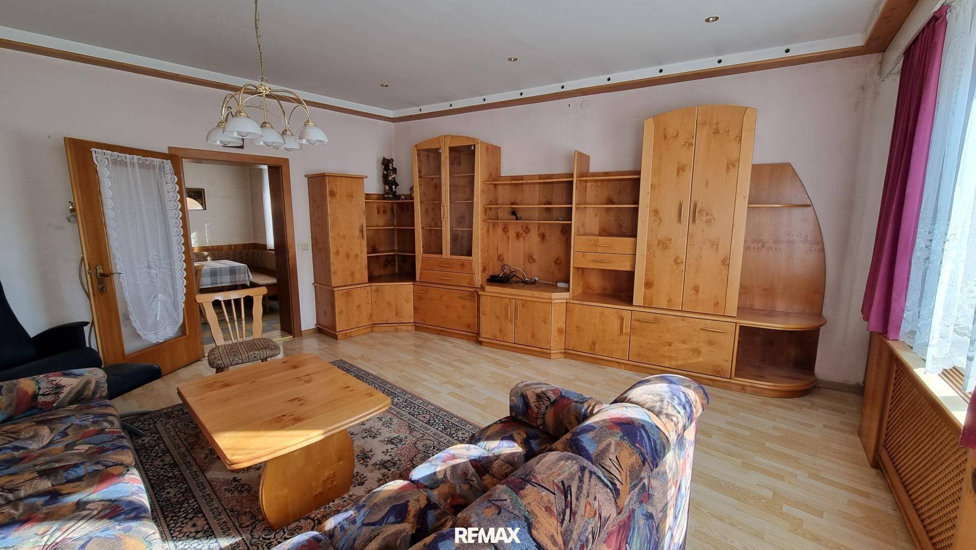 Wohnzimmer mit Holzmöbeln, gemustertem Sofa und Blick in einen angrenzenden Essbereich.