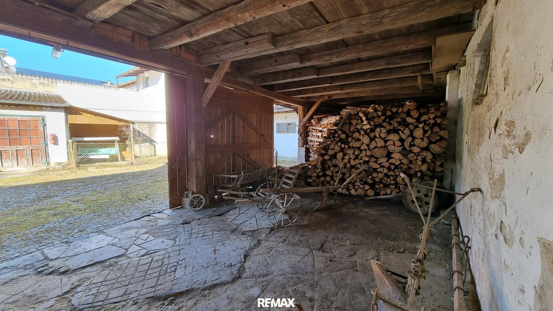 Offene Scheune mit gestapeltem Brennholz, Holzbalken und alten landwirtschaftlichen Geräten.
